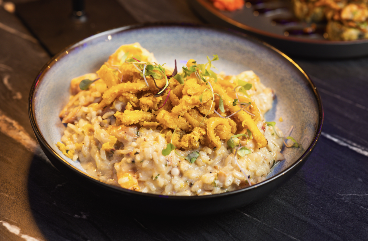 Bowl of creamy seafood risotto topped with fried calamari and microgreens on a black plate.