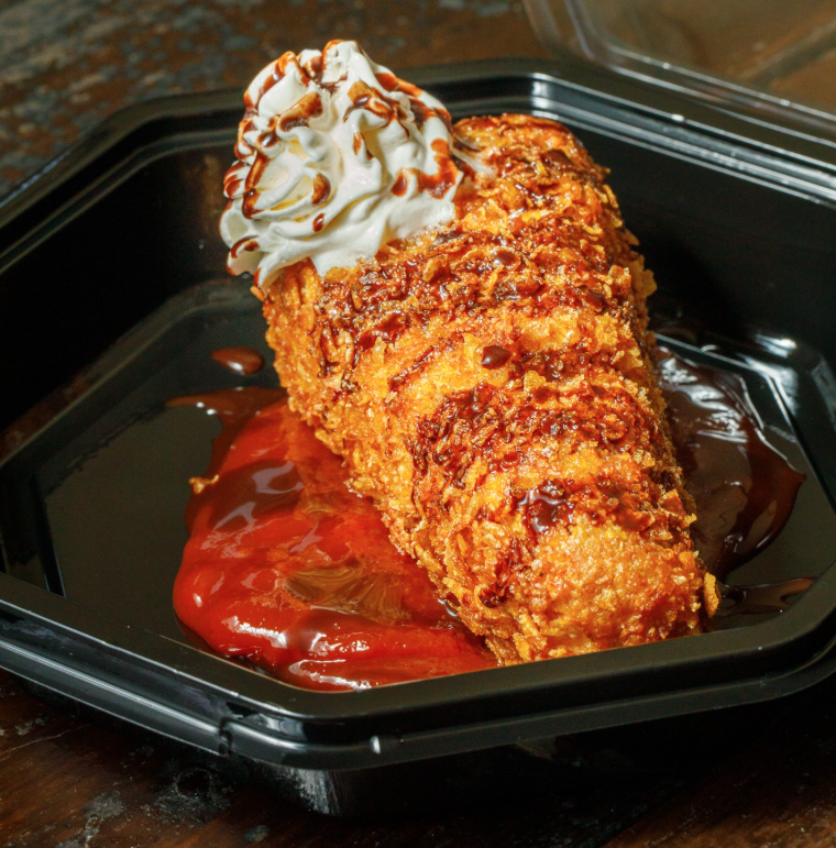 Fried chicken breast topped with whipped cream and drizzled with caramel sauce, served with ketchup in a black takeout container.