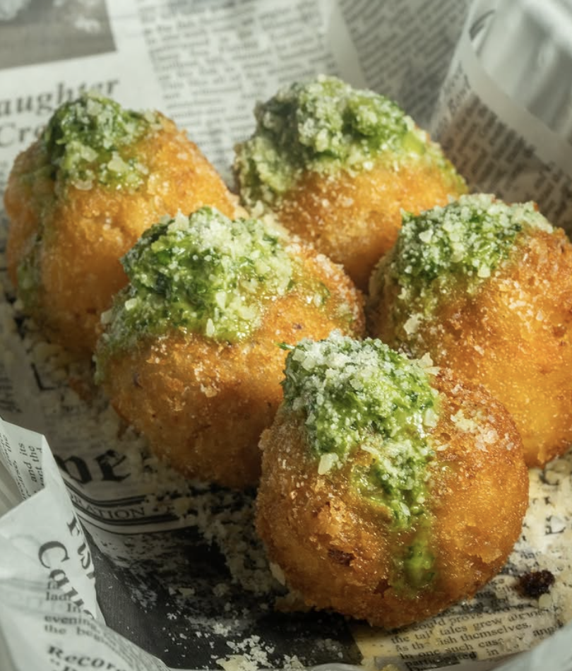 Fried cheese balls topped with green sauce and grated cheese, served on newspaper-style paper