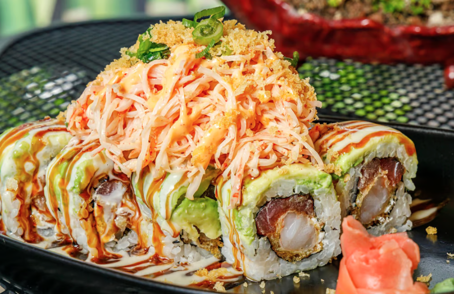 Close-up of a sushi roll topped with shredded crab meat, green onions, spicy mayo, and eel sauce, served with pickled ginger on the side.