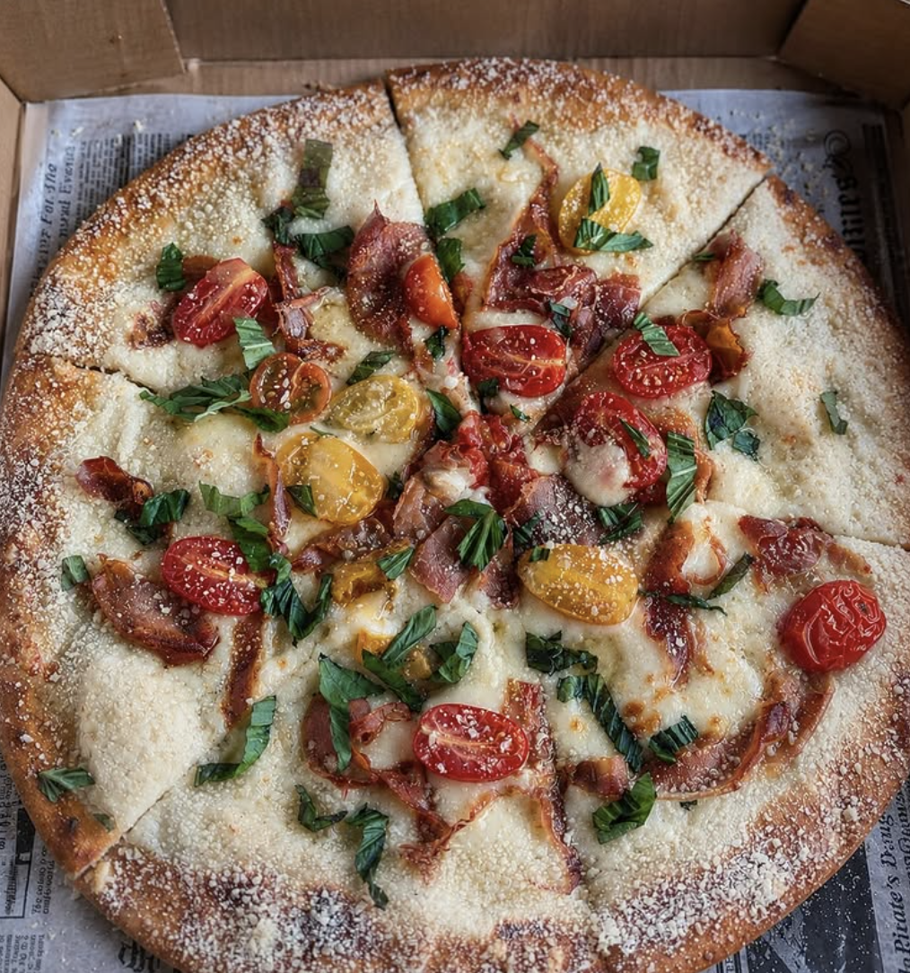 A pizza with a golden crust topped with cherry tomatoes, bacon, chopped green herbs, and grated cheese.
