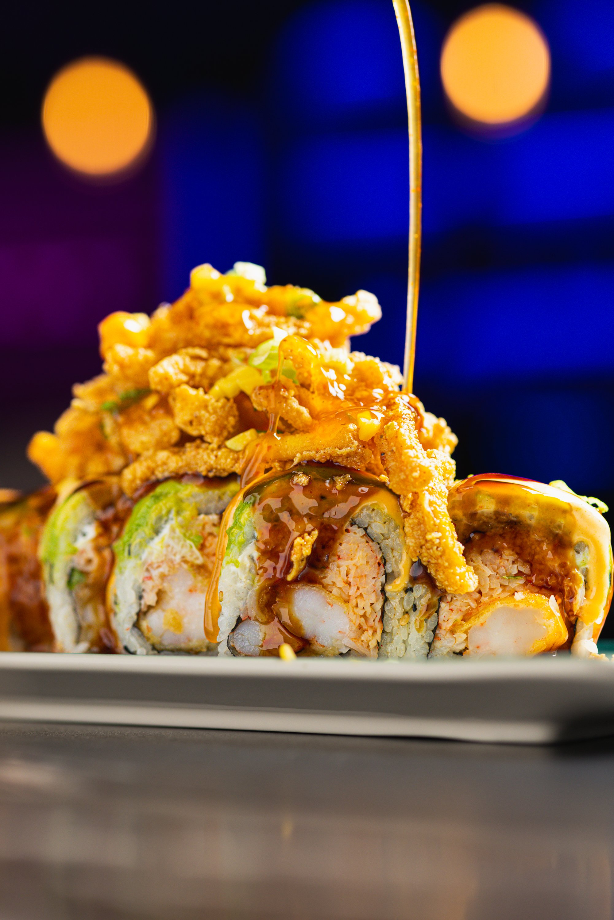 Close-up of a sushi roll topped with crispy tempura flakes, eel sauce, and thin slices of green avocado on a white plate with colorful bokeh lights in the background.