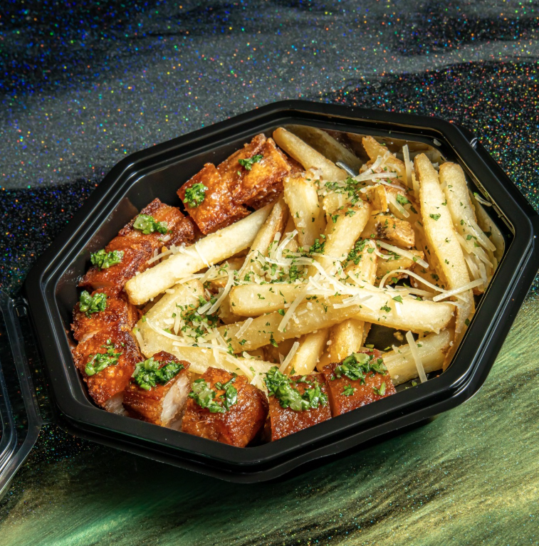 Black container holding fried chicken bites with green herbs, French fries topped with cheese and herbs, garnished with parsley, on a glittery surface.