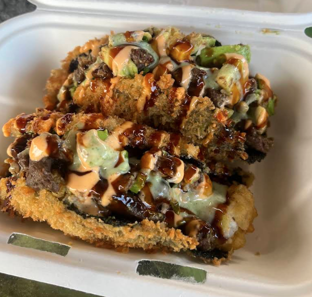A close-up of a piece of spicy sushi roll topped with eel sauce, spicy mayo, crunchy tempura bits, chopped green onions, and avocado, served in a white takeout container.
