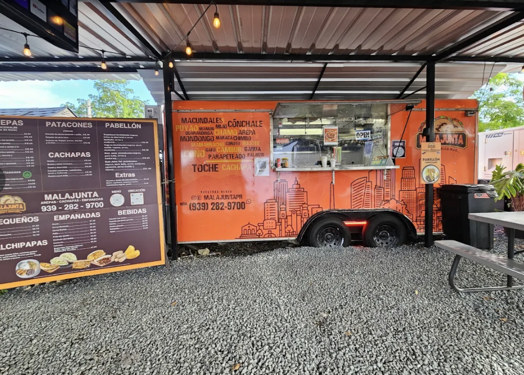 Food truck with orange exterior selling Mexican snacks, with a menu sign on the left listing various items, parked under a shelter with string lights, set on gravel ground.