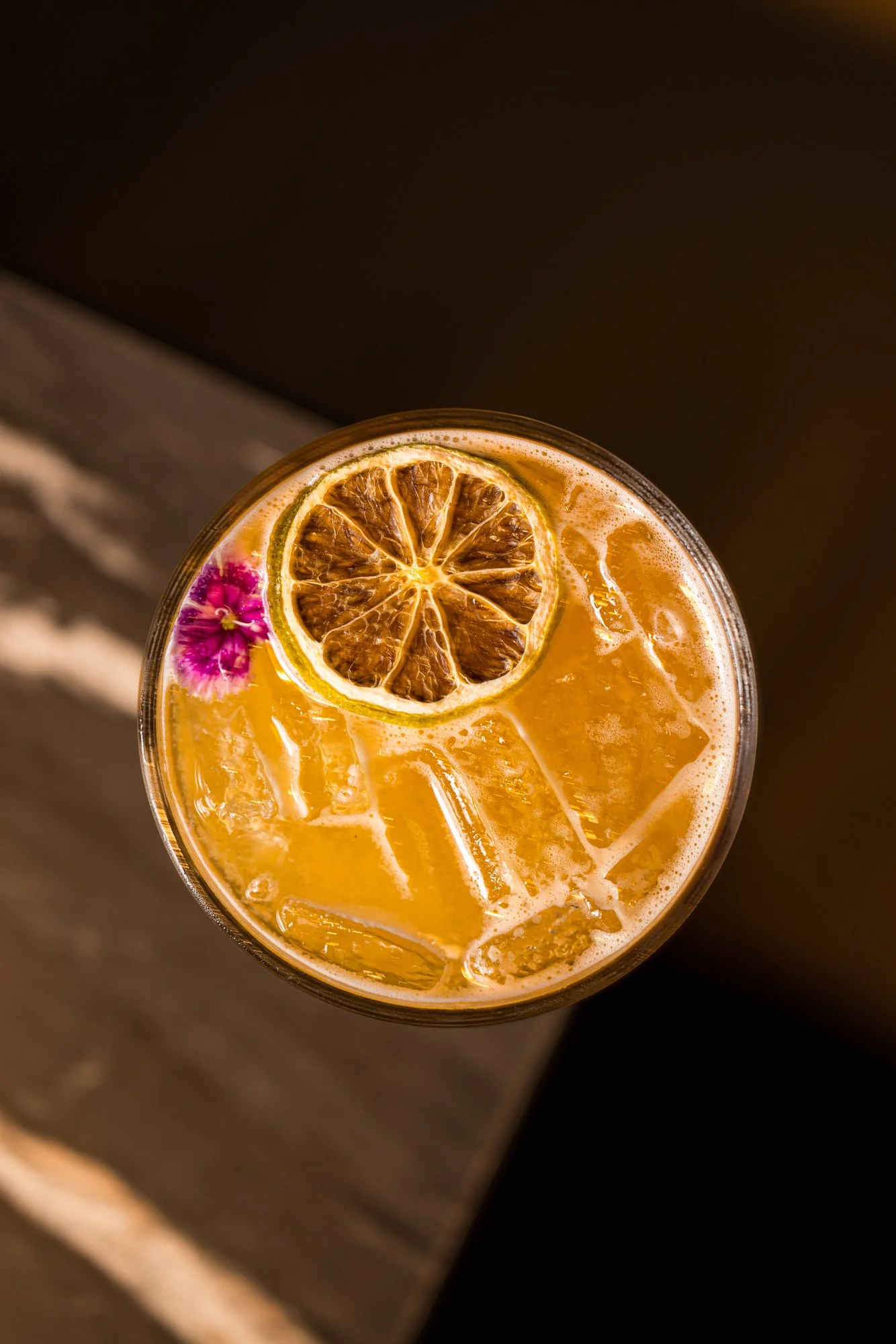 A top view of a glass of orange-colored drink with ice, garnished with a dried lemon slice and a purple flower.