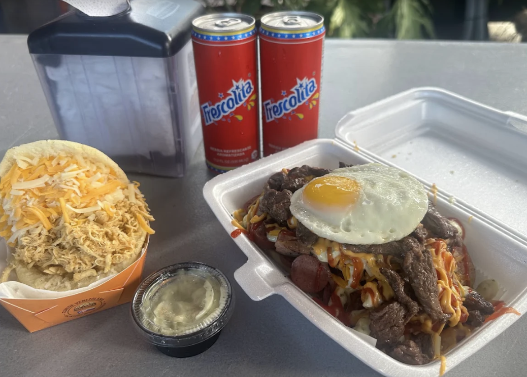 A takeout meal with a beef and noodles dish topped with a fried egg, a sandwich with shredded chicken and cheese, two cans of Frescolita soda, a small side of sauce or sour cream, and a napkin holder on a table outdoors.