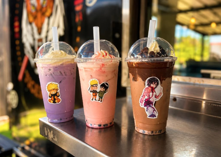 Three different flavored bubble teas in clear cups with cartoon character stickers, each with a straw and plastic dome lid, placed on a metal surface outdoors.