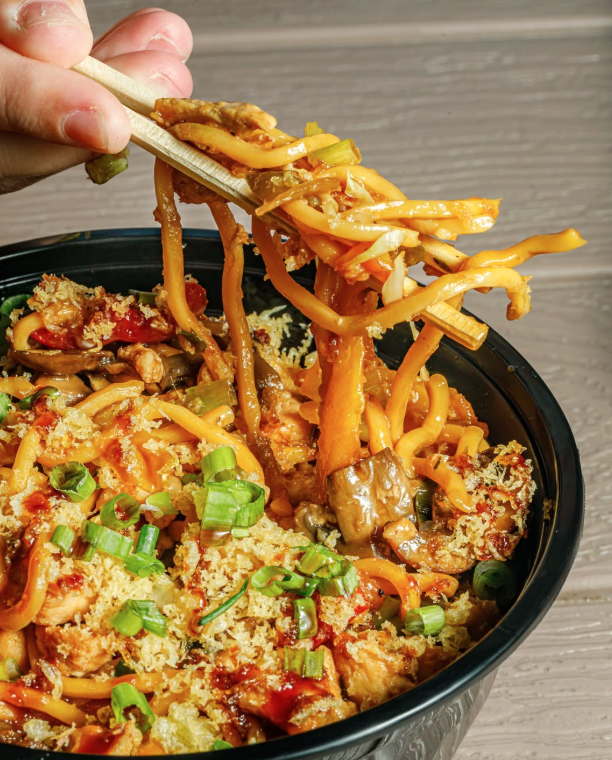 Close-up of a black bowl of chicken and vegetable stir fry topped with green onions, with chopsticks lifting some noodles.