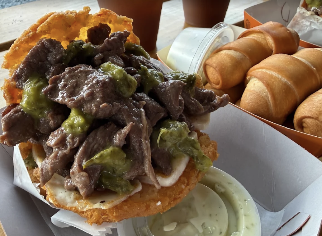 A cheeseburger topped with shredded beef and green pepper slices, with a side of fried taquitos and dipping sauce.