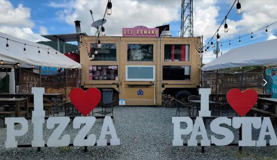 Outdoor pizza and pasta food stand decorated with large white letters spelling 'I ❤️ PIZZA' and 'I ❤️ PASTA,' surrounded by string lights, tents, and gravel ground.
