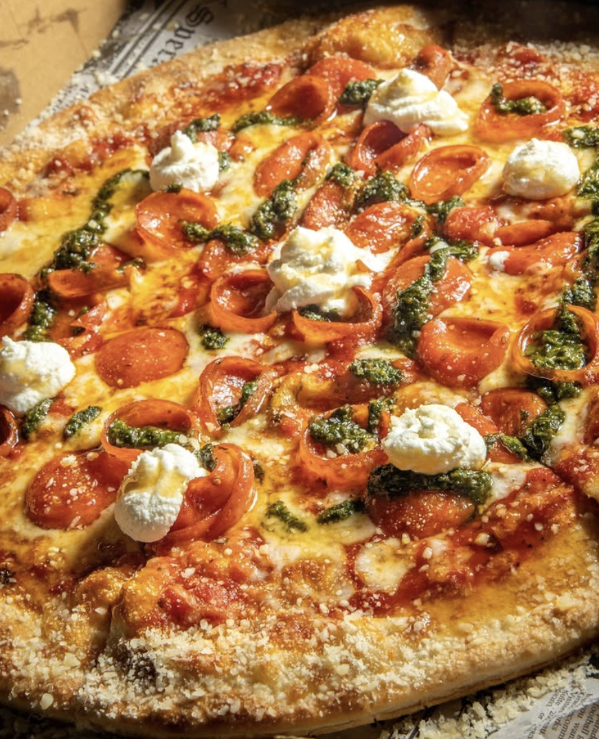 Close-up of a pepperoni pizza with cheese, dollops of ricotta cheese, and green pesto sauce on top.