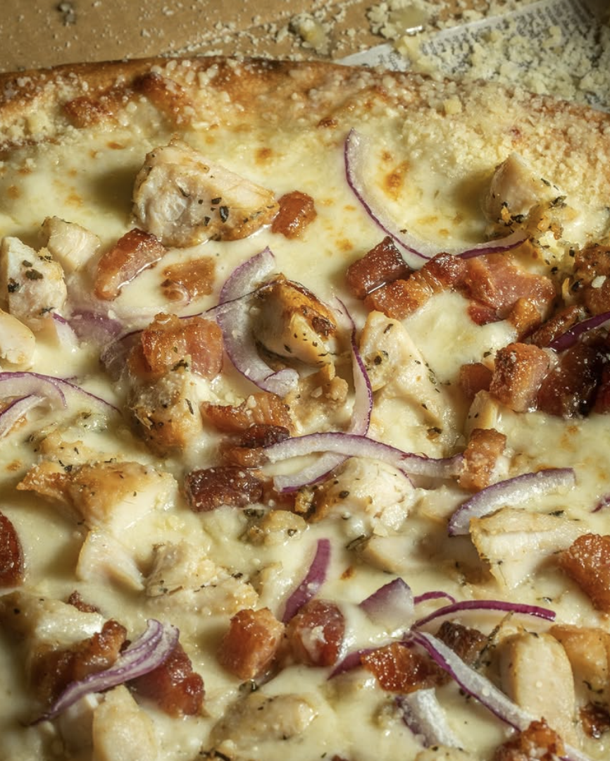 Close-up of a pizza with cheese, chicken pieces, bacon bits, and sliced red onions on a pizza crust.