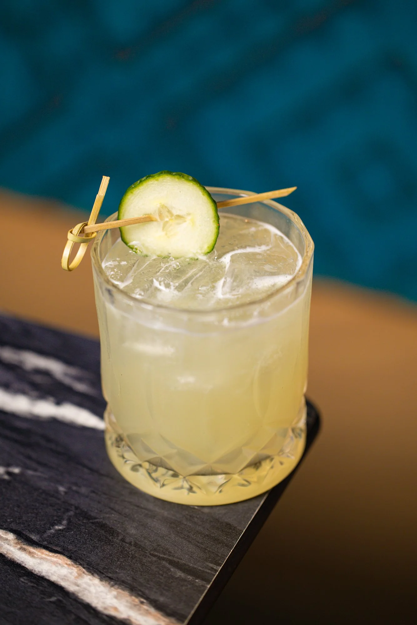 A glass of lemonade garnished with a cucumber slice and a decorative skewer, placed on a black slate board.