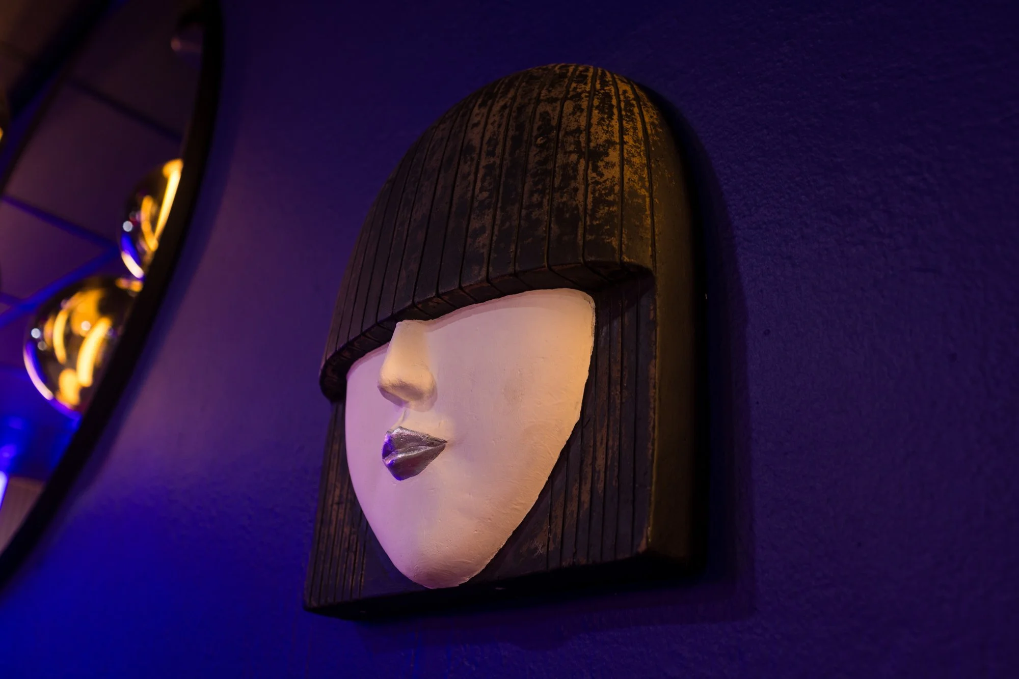 Decorative wall art featuring a Woman's face with closed eyes, metallic lips, and a beanie hat, illuminated with purple and warm lighting.