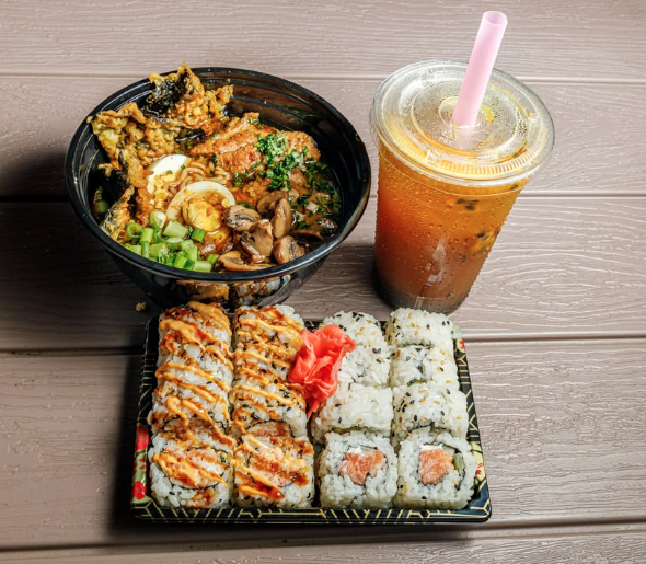 A tray with sushi rolls, pickled ginger, and wasabi, a bowl of ramen with boiled eggs, vegetables, and broth, and a plastic cup of iced tea with a straw on a wooden table.