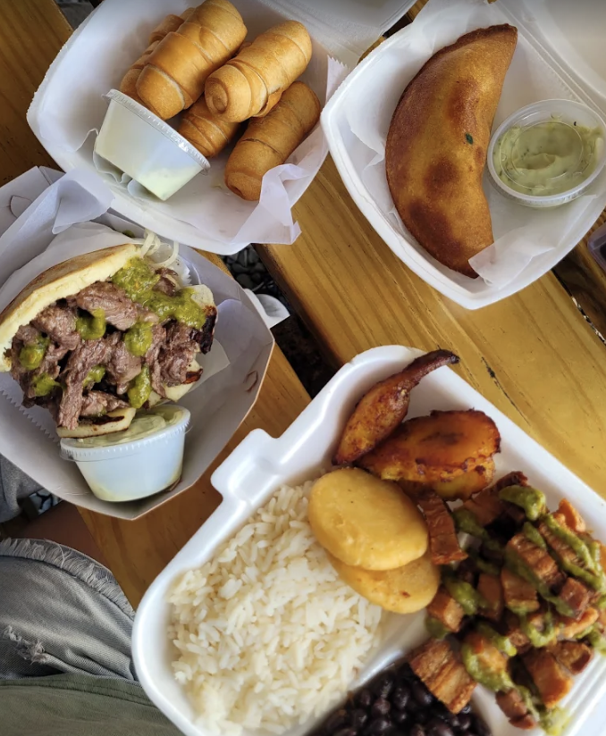 Various takeout foods including breaded fried items, a cheeseburger with green relish, rice, beans, grilled meat with sauce, fried plantains, and a calzone with dipping sauce on a wooden table.