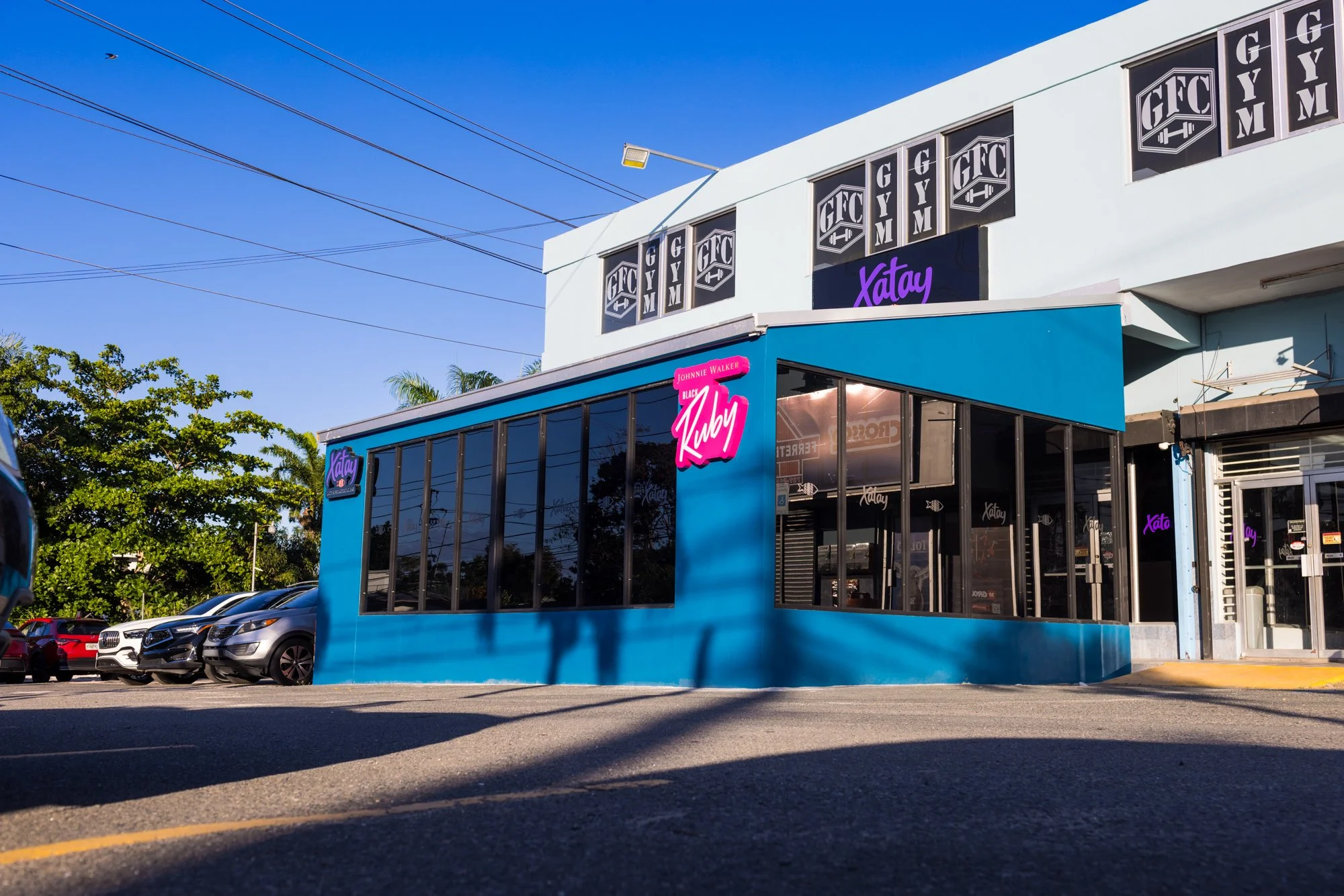A fitness gym building with signage reading 'GFC GYM' on the upper windows and 'Xatay' on a black sign. The building has large windows and a blue exterior. There are parked cars along the street and some green trees nearby, with a clear blue sky overhead.