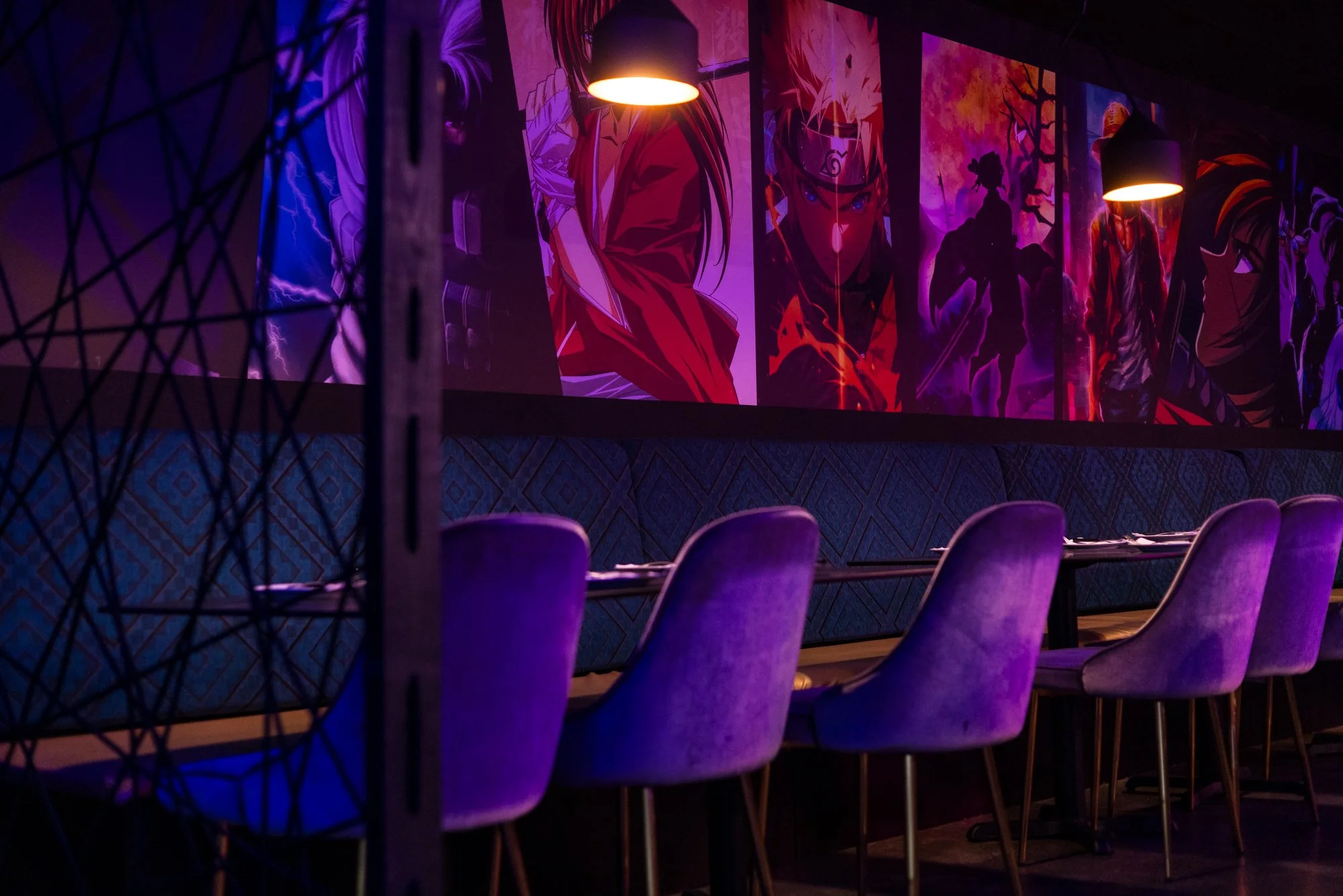 Dimly lit room with purple chairs and a long table, decorated with Naruto-themed posters and artwork on the wall illuminated by warm lighting.