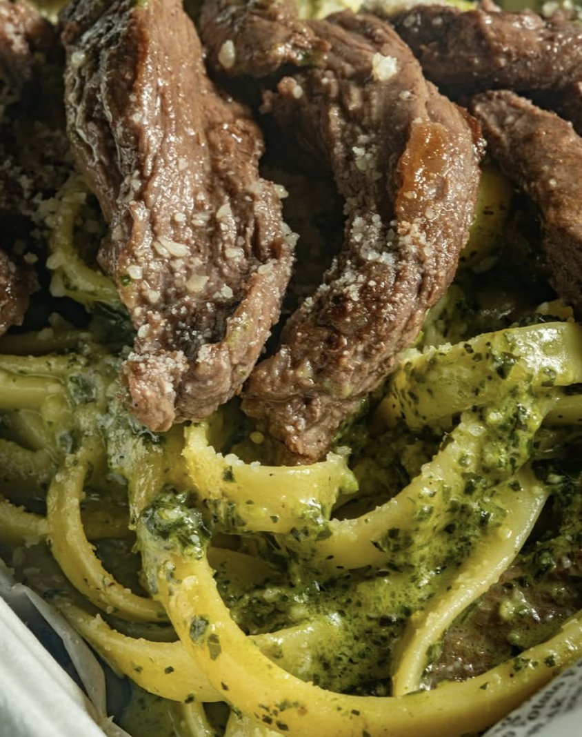 Close-up of cooked pasta with green herbs, topped with cooked beef strips and grated cheese.