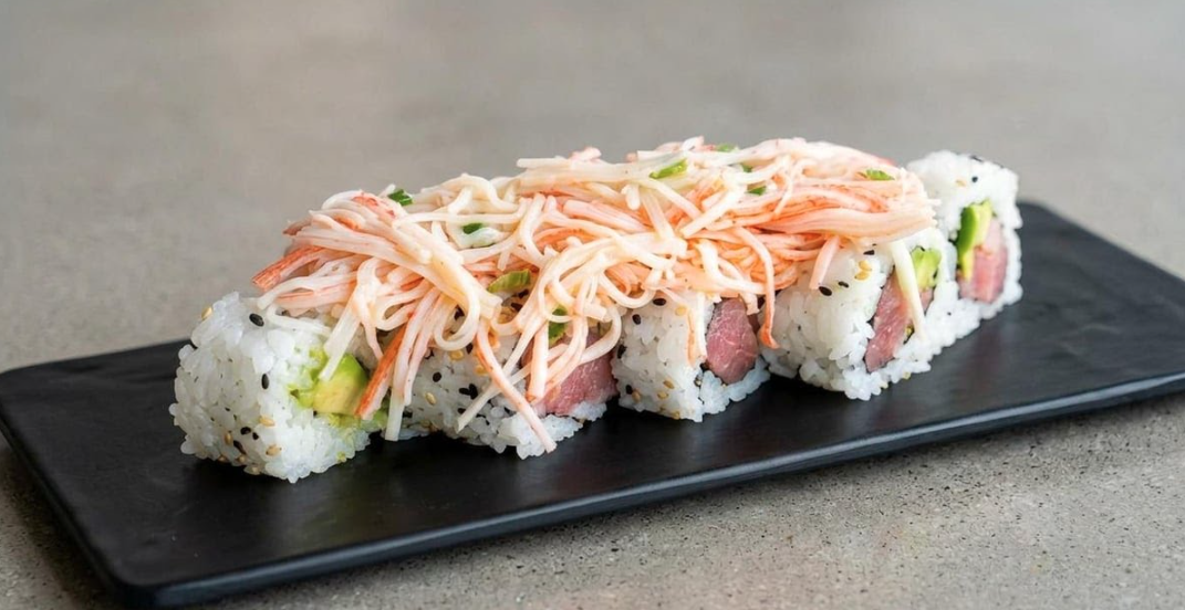 Close-up of a sushi roll with rice, raw fish, avocado, topped with crab meat and shredded crab salad on a black rectangular plate.
