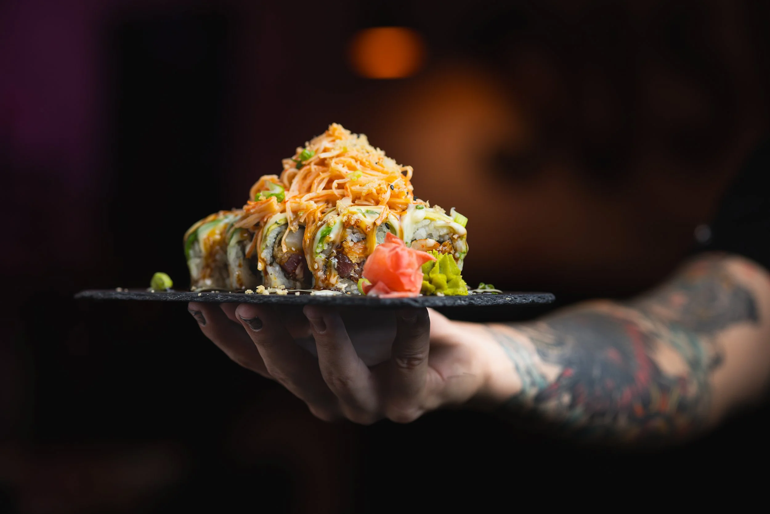 Person holding a black tray with a colorful sushi roll topped with shredded salmon, green onions, and drizzled sauce, against a dark blurred background.