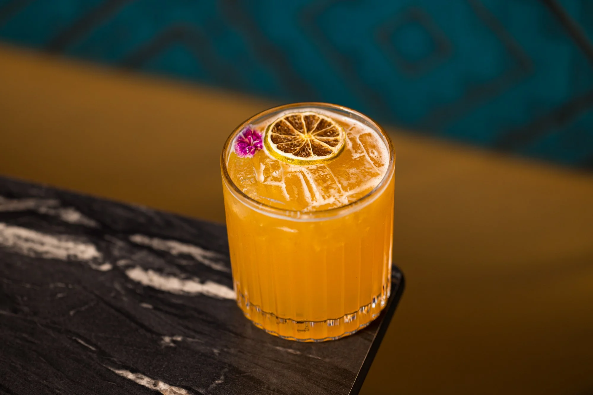 A glass of orange-colored beverage garnished with a slice of dried lemon and a small pink flower on top, placed on a dark wooden surface.
