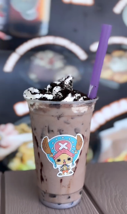 Chocolate milkshake topped with whipped cream and crushed cookies, with a purple straw, in a clear cup with a cartoon sticker, on a table with a blurred dessert menu in the background.