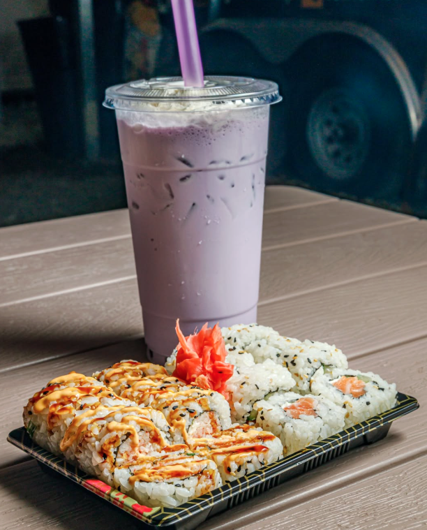 Sushi rolls with spicy salmon, a dollop of pink ginger, and white rice with black sesame seeds, accompanied by a large iced pink milk drink with a purple straw, all on a black rectangular plate on a wooden table.