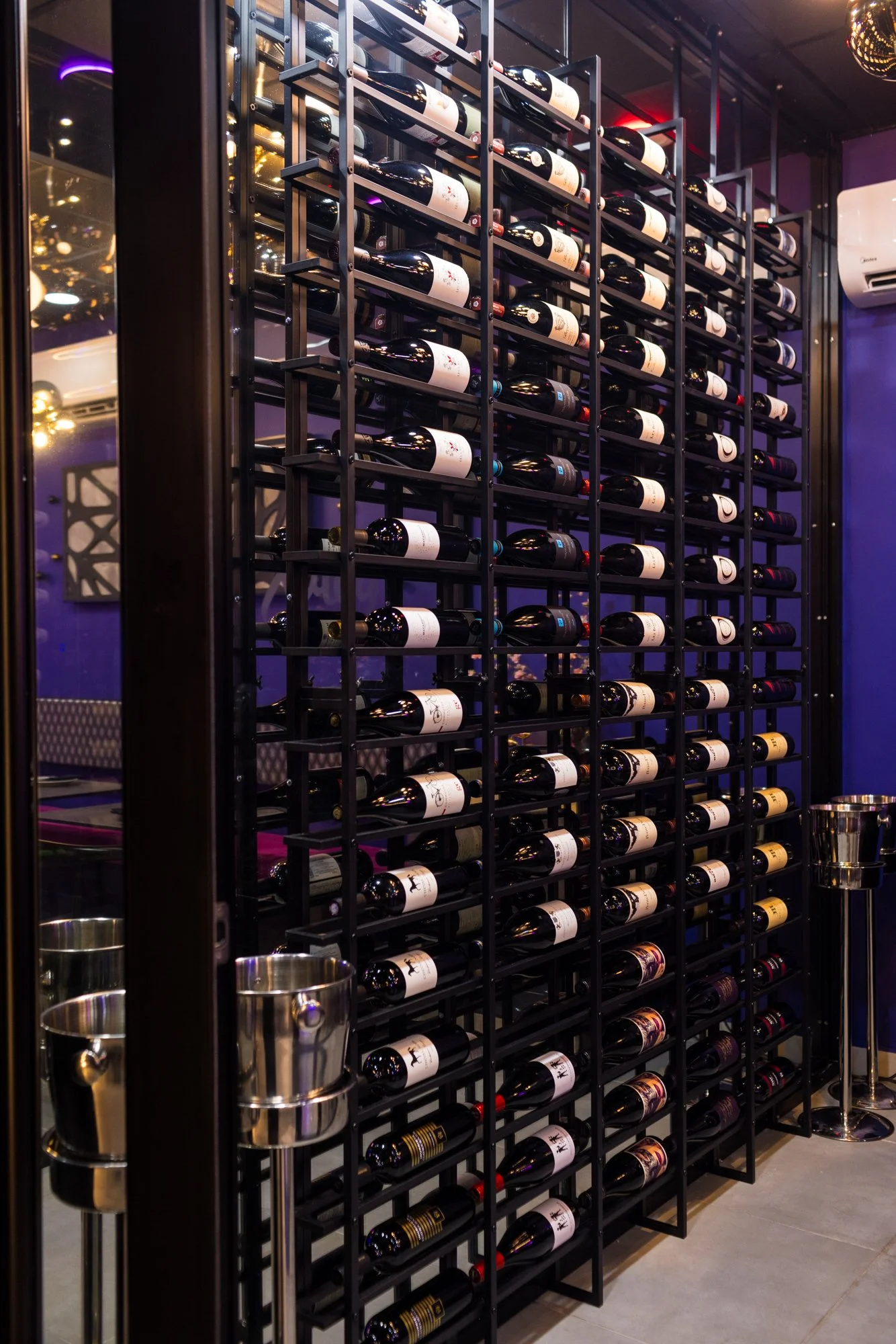 A wine storage rack filled with bottles in a restaurant or bar setting. The rack is black metal with multiple shelves, each holding several bottles of wine. There are some metallic wine coolers hanging on the rack, and the room has purple and yellow 