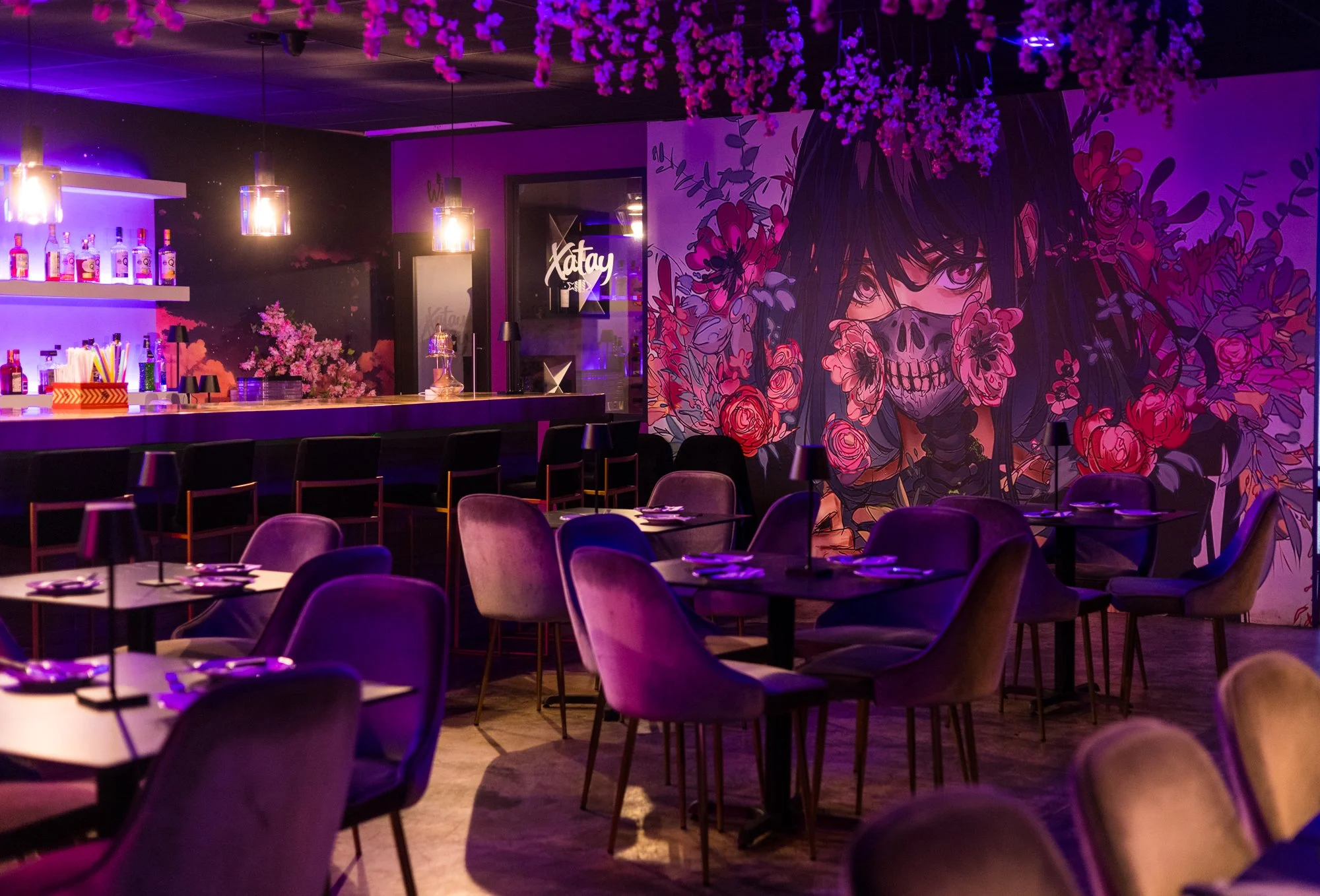 An interior view of a dimly lit bar or lounge area with purple and pink lighting. There are tables with chairs, a bar counter with bottles and mixers, and a large artistic mural of a girl with flowers and a skull mask on the wall.