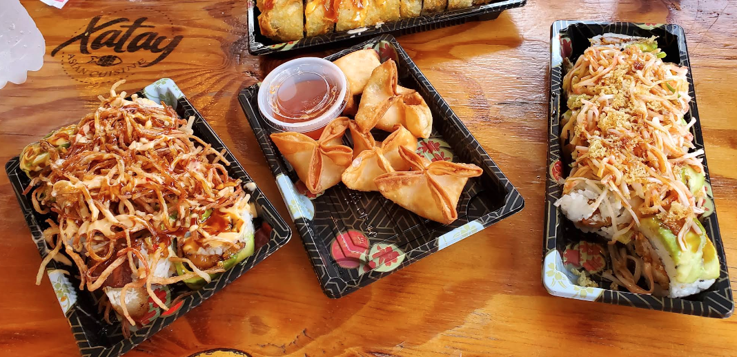 Three black trays of assorted sushi and spring rolls on a wooden table, with dipping sauce in a small cup in the middle.