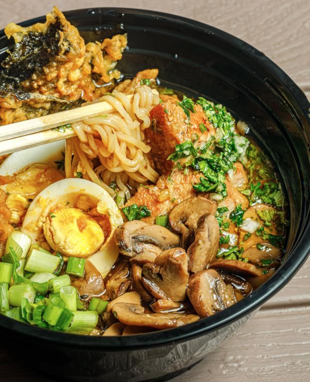 A black bowl of ramen with sliced mushrooms, halved boiled egg, chopped green onions, vegetable tempura, and meatballs garnished with chopped herbs.