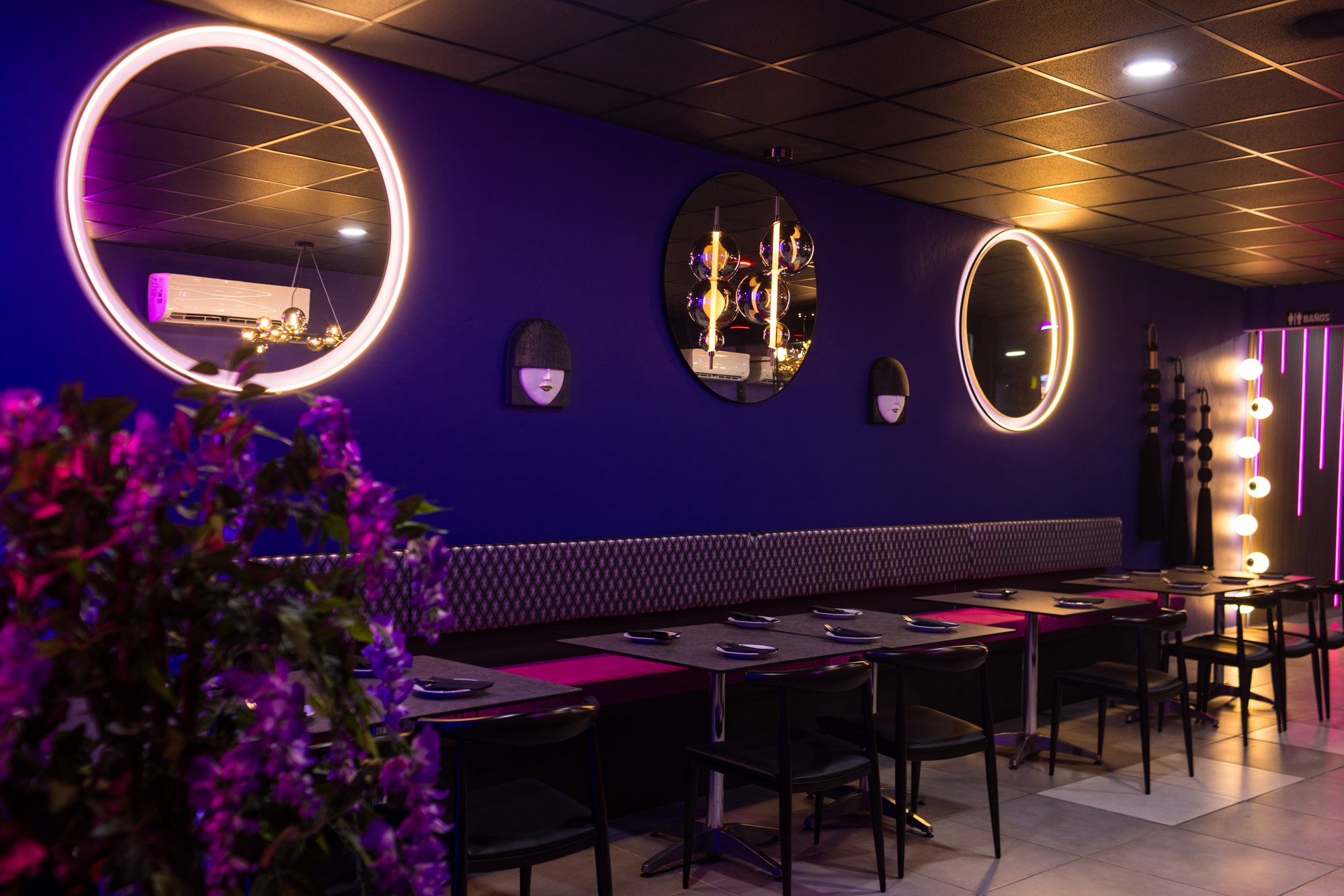 A modern restaurant interior with purple walls, bright neon circular lights, decorative hanging lamps, wall art with human faces, and a row of tables set with plates and cutlery. Purple flowers are visible in the foreground.