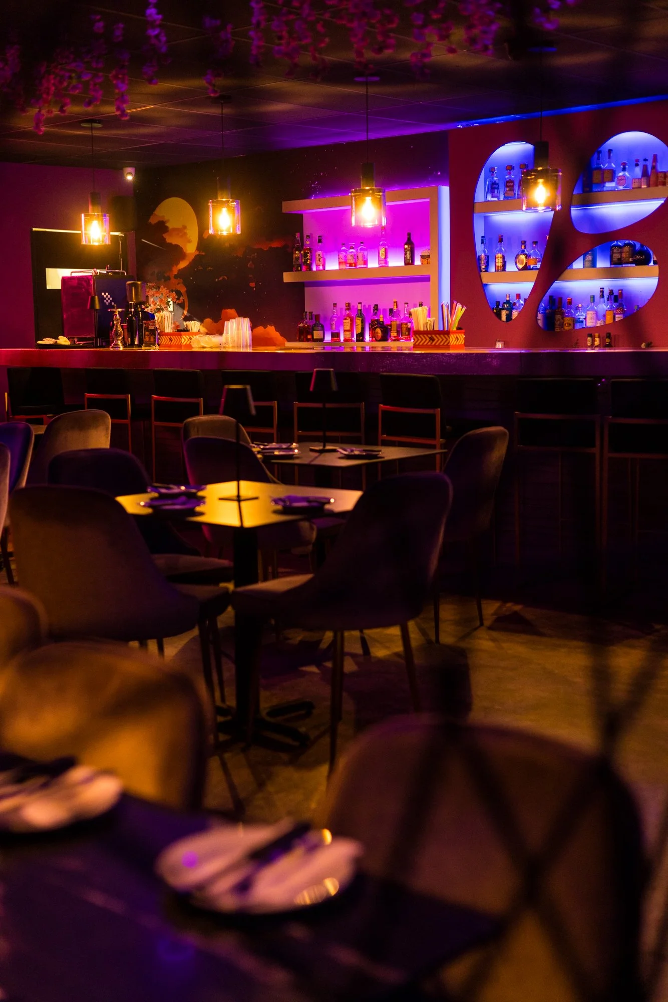 A dimly lit bar with purple and blue lighting, shelves with various bottles of alcohol, a bar counter with glasses and bar tools, and tables with chairs, some with napkins and place settings, and a person in the foreground holding a plate.