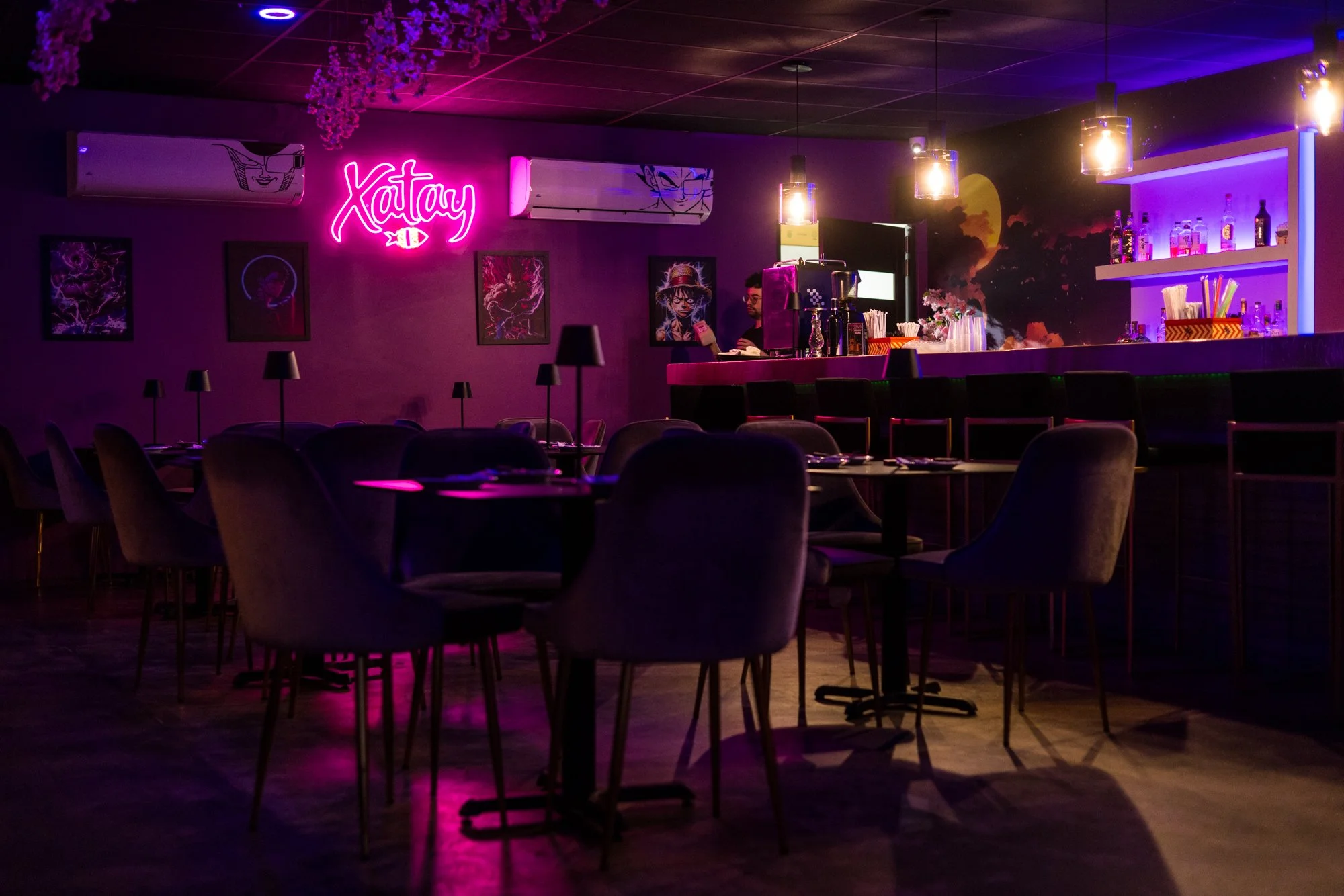 A dimly lit bar with purple and pink neon lighting. There are several tables with chairs, dark lamps, and colorful artwork on the wall. The bar counter displays various bottles and bar supplies. A bartender is working behind the counter.