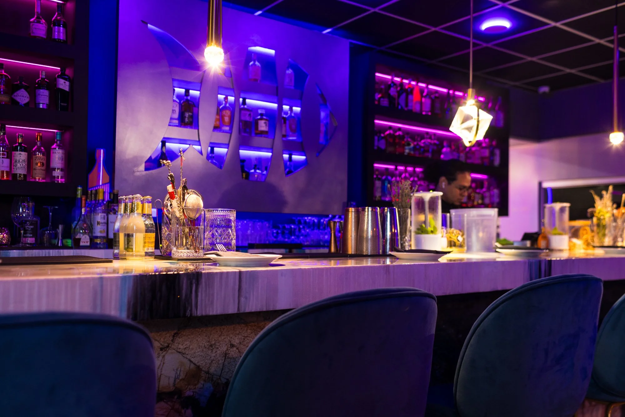 A bar with purple and blue neon lighting, bottles of alcohol on shelves, a bartending station with glasses and drinks, and a bartender preparing drinks.