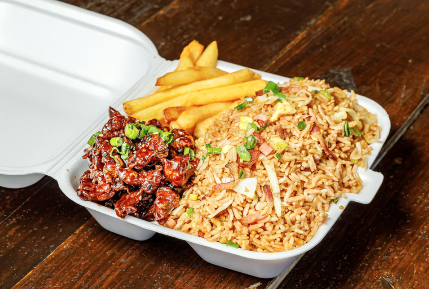 A takeout container with fried rice, French fries, and glazed teriyaki chicken garnished with chopped green onions, placed on a wooden table.