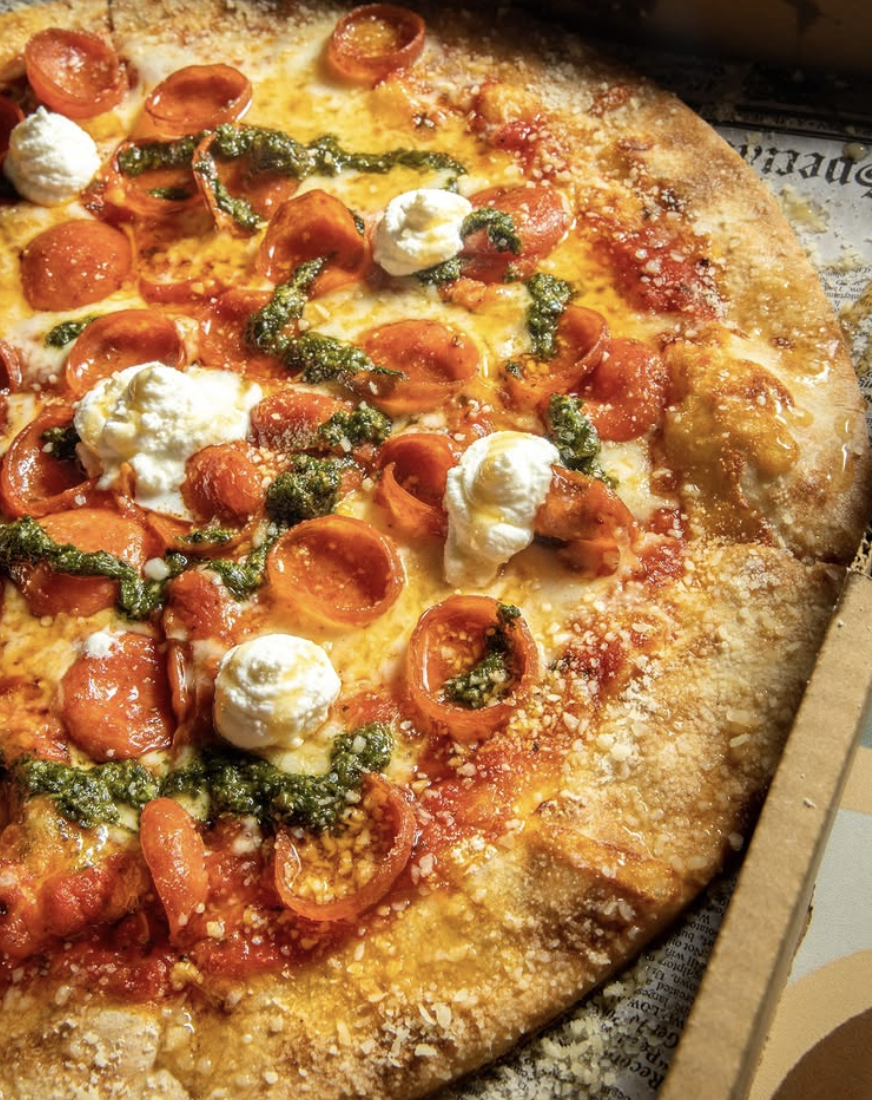 Close-up of a pepperoni and cheese pizza with dollops of ricotta cheese, green pesto, and a sprinkle of grated Parmesan cheese.