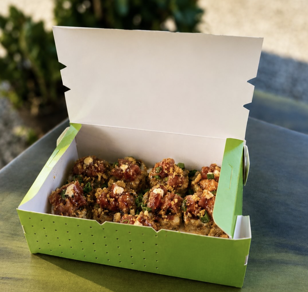 A green and white takeout box filled with sushi topped with chopped green onions, sesame seeds, and sauce, set on a dark surface outdoors.