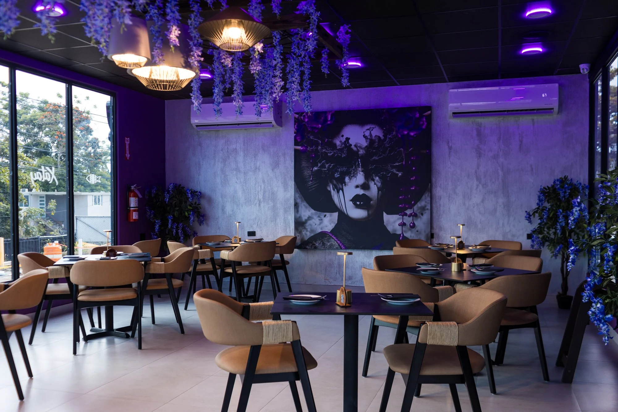 Interior of a stylish modern restaurant with beige chairs, black tables set with napkins and tableware, purple LED ceiling lights, a large black and white artwork of a woman's face with artistic effects, purple floral decorations hanging from the cei