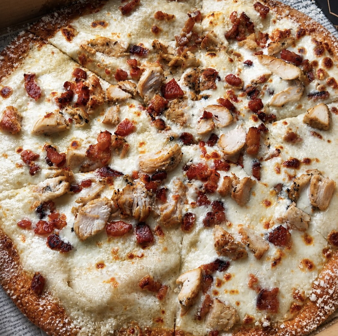 Close-up of a pizza topped with cheese, grilled chicken pieces, chopped tomatoes, and bacon bits.