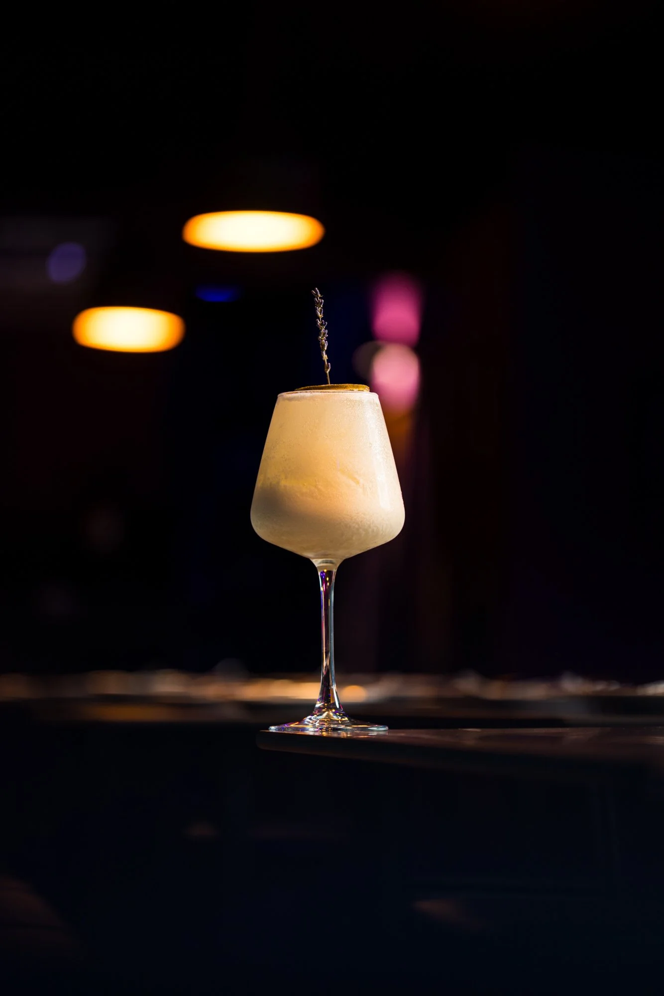 A cocktail glass filled with a creamy drink garnished with a sprig of lavender, set against a dark background with blurred colorful lights.