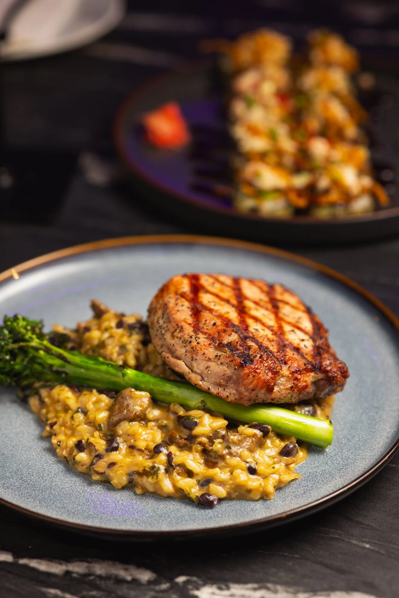 A grilled chicken breast served with a side of creamy black bean and corn mixture and a cooked green vegetable on a gray plate. In the background, a skillet with a vegetable stir-fry or salad is visible.