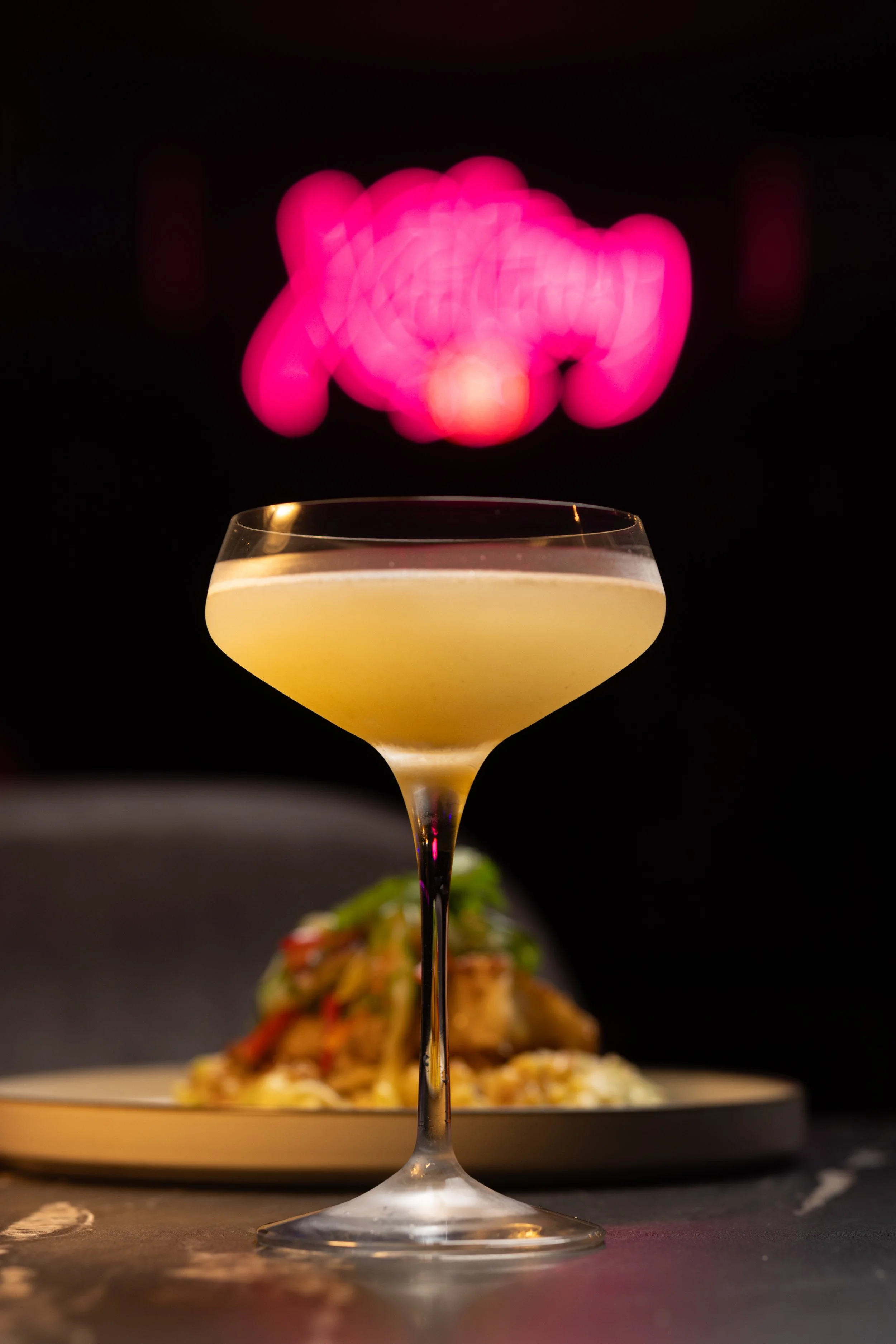 A cocktail glass filled with a yellow drink, with a blurred plate of pasta dish in the background, and vibrant pink and red bokeh lights behind the glass.