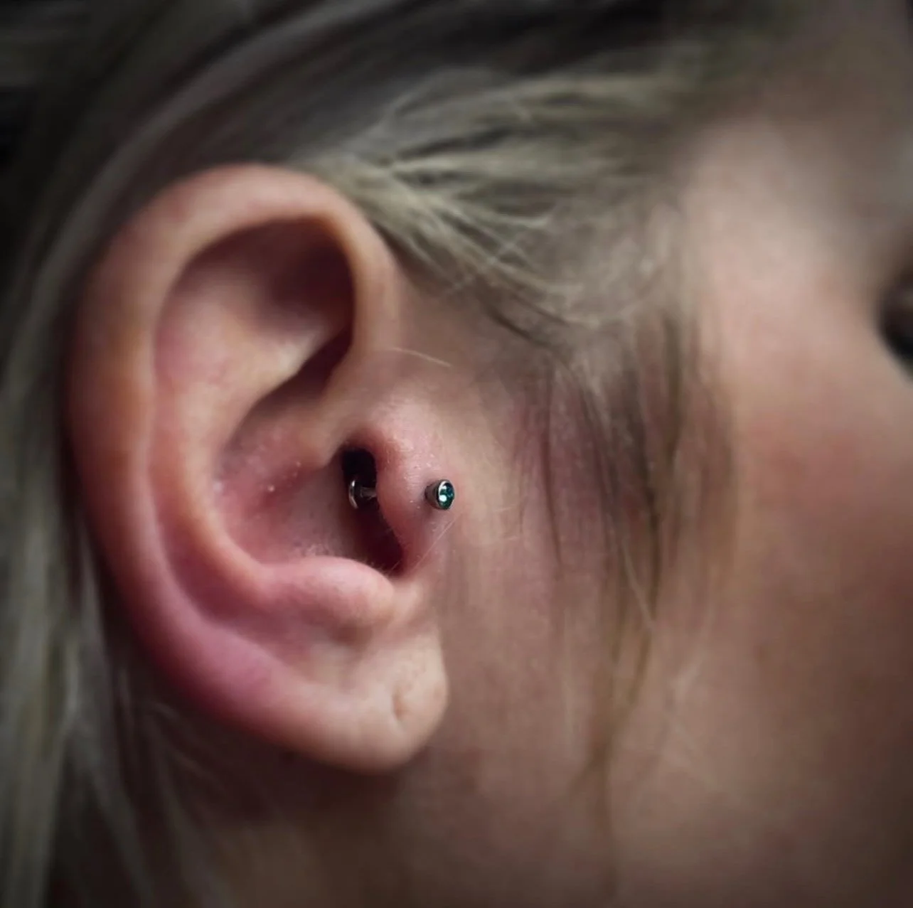 Close-up of a person's ear with a tongue piercing featuring a black stud, with blonde hair and skin visible.