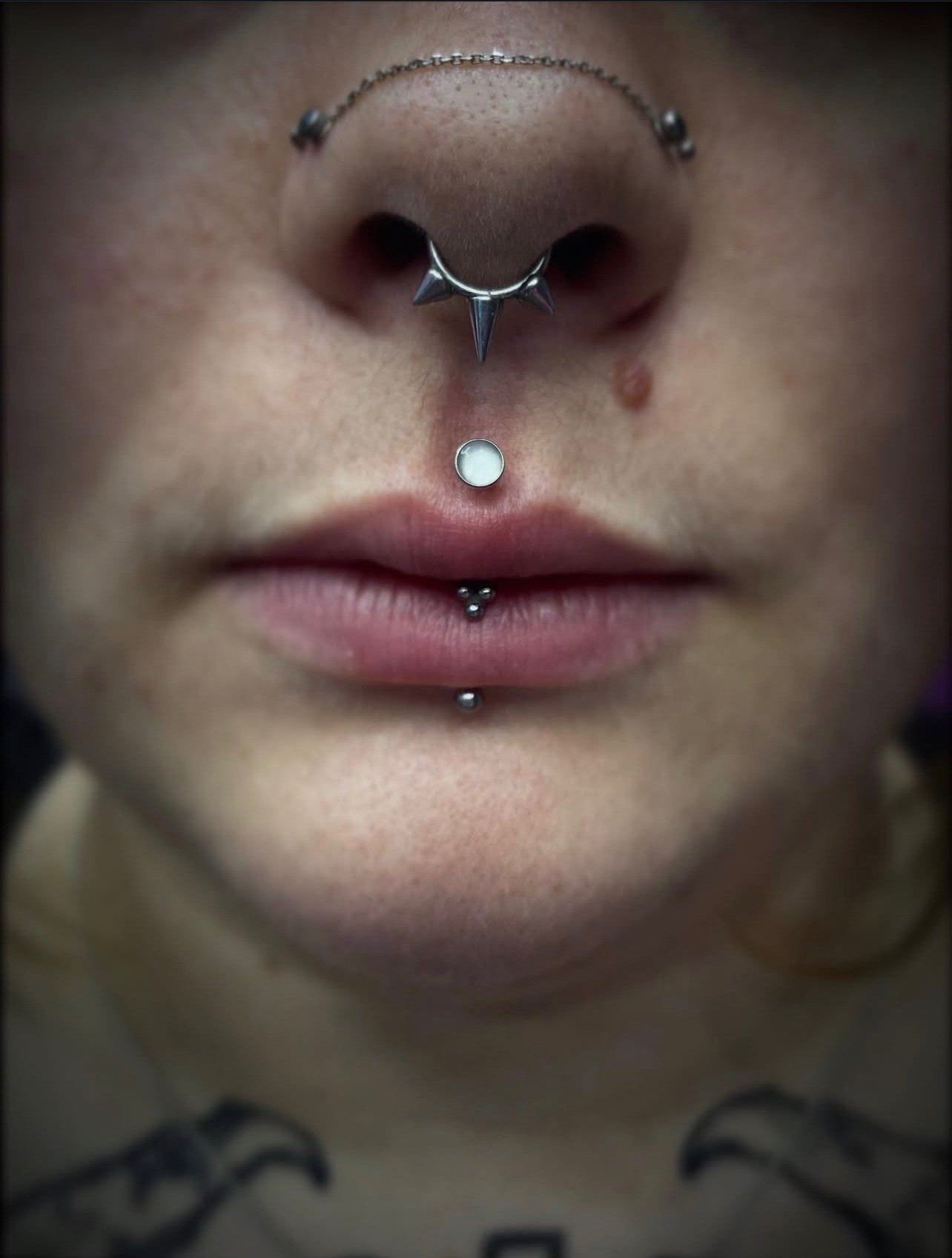 Close-up of a person's nose and lips, featuring multiple facial piercings, including a septum ring with spikes, a nostril piercing, and a labret and tongue piercing, with tattoos visible on the neck.