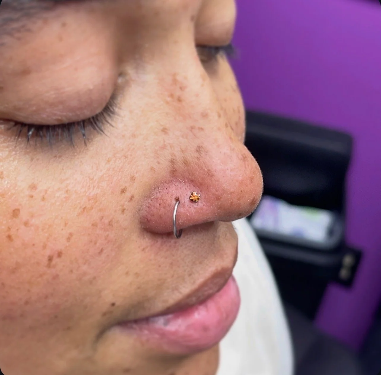 Close-up of a person's face showing freckles, closed eyes, and a nose piercing with a small gold stud.