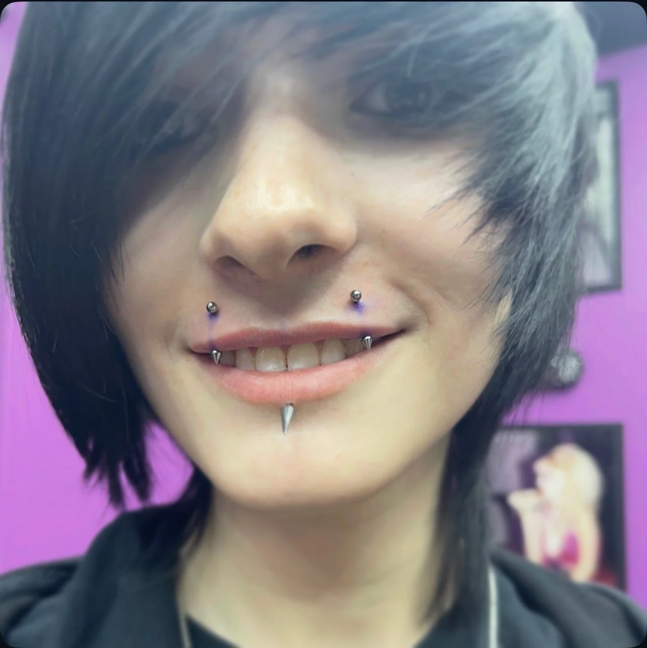 A person with black, layered hair and multiple facial piercings, including a septum piercing and a labret piercing, smiling with visible teeth, against a purple background.