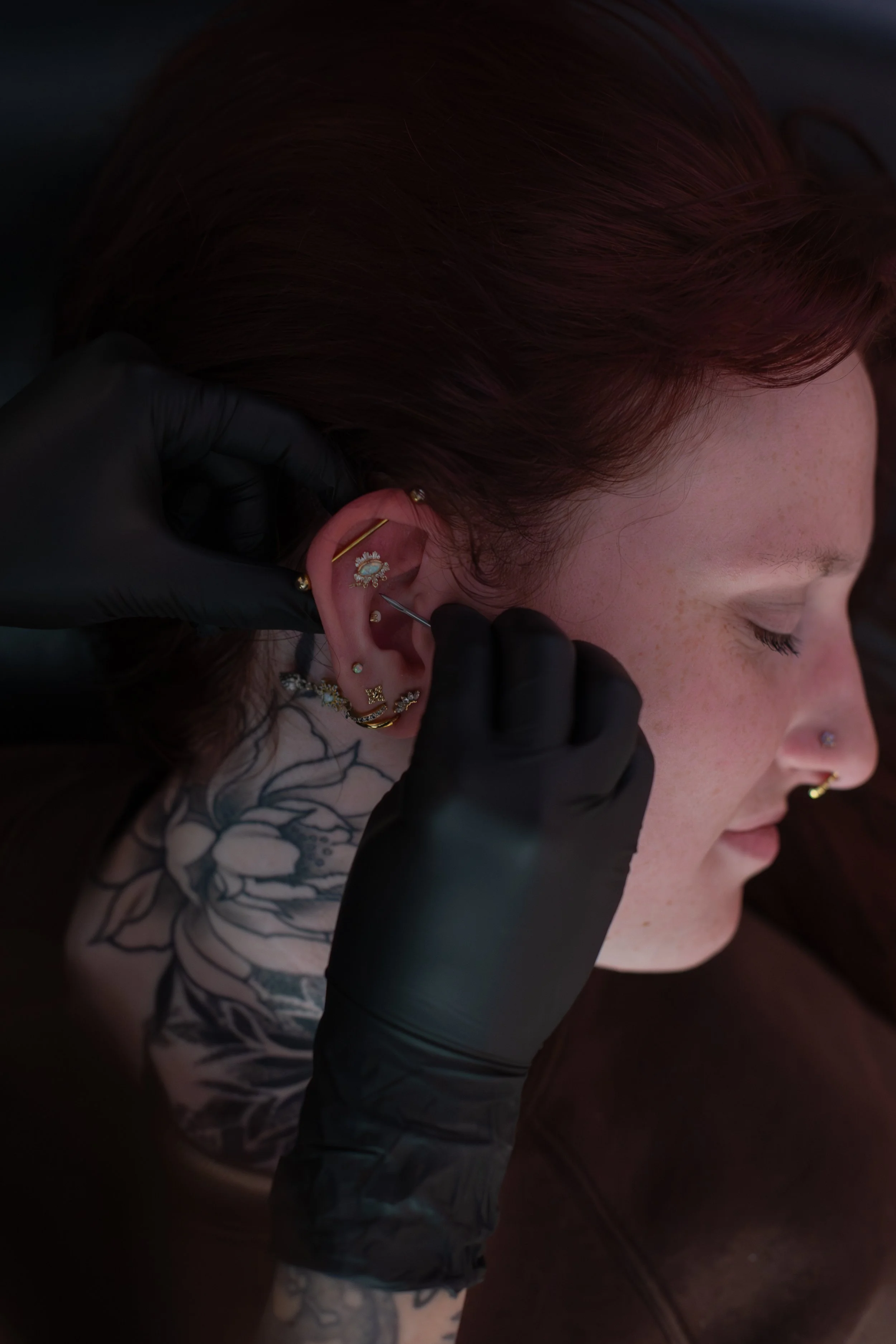 A person with red hair has multiple earrings pierced in their ear, and a tattoo on their arm. A gloved hand is inserting or adjusting an earring in their ear.